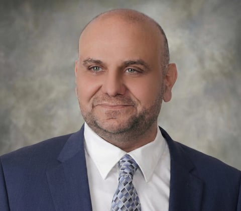 Professional headshot of a bald man with beard wearing a dark blue suit, white shirt, and blue patterned tie against a gray background